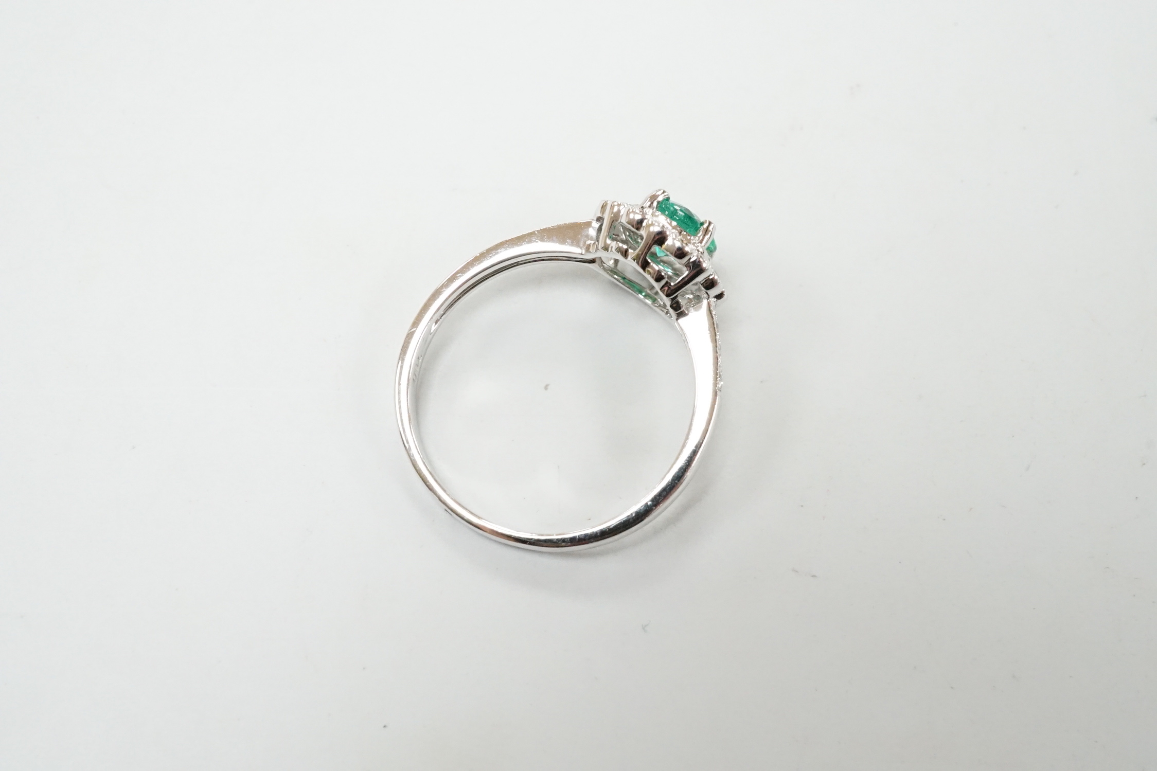 A modern 18ct white gold, emerald and diamond set circular cluster ring, with diamond set shoulders, size M, gross weight 2.7 grams.